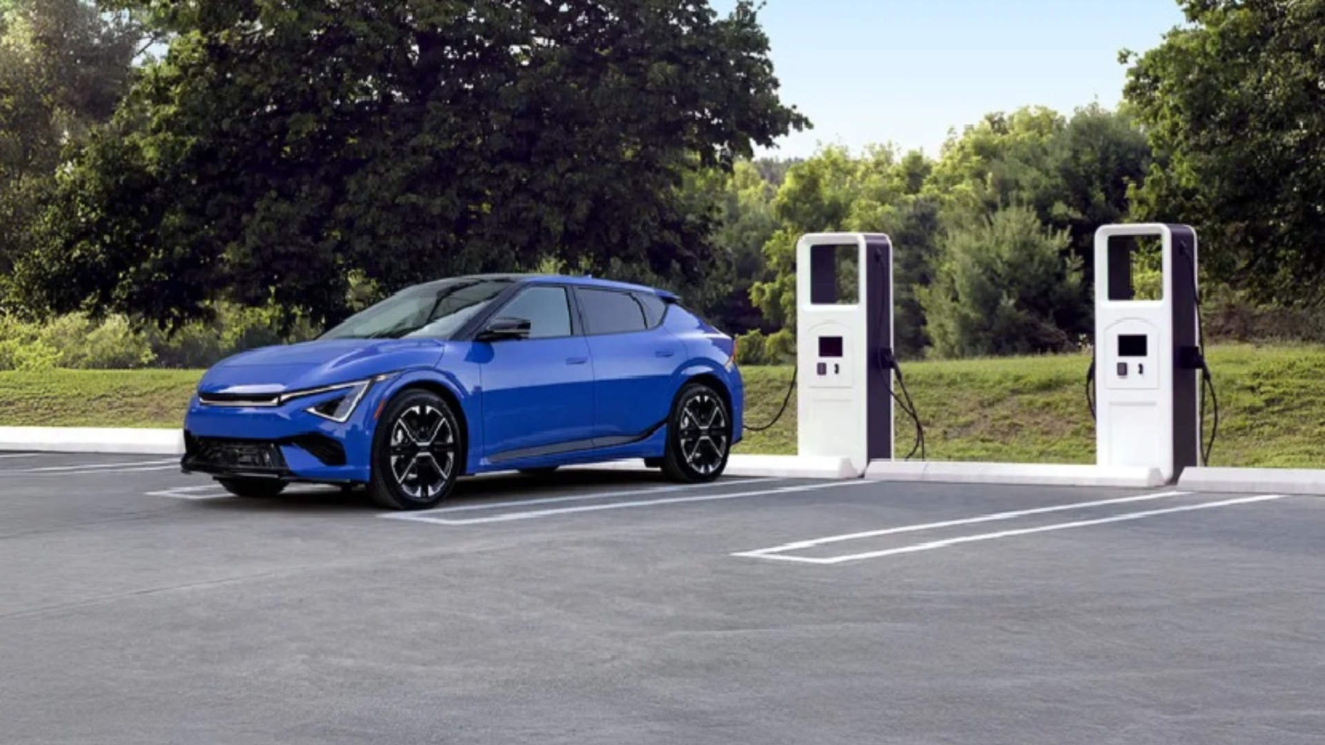 Blue electric vehicle charging at two white fast-charging stations in outdoor parking area with green trees