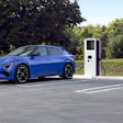 Blue electric vehicle charging at two white fast-charging stations in outdoor parking area with green trees