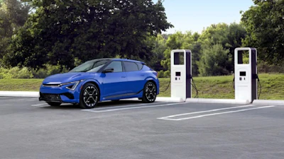Blue electric vehicle charging at two white fast-charging stations in outdoor parking area with green trees