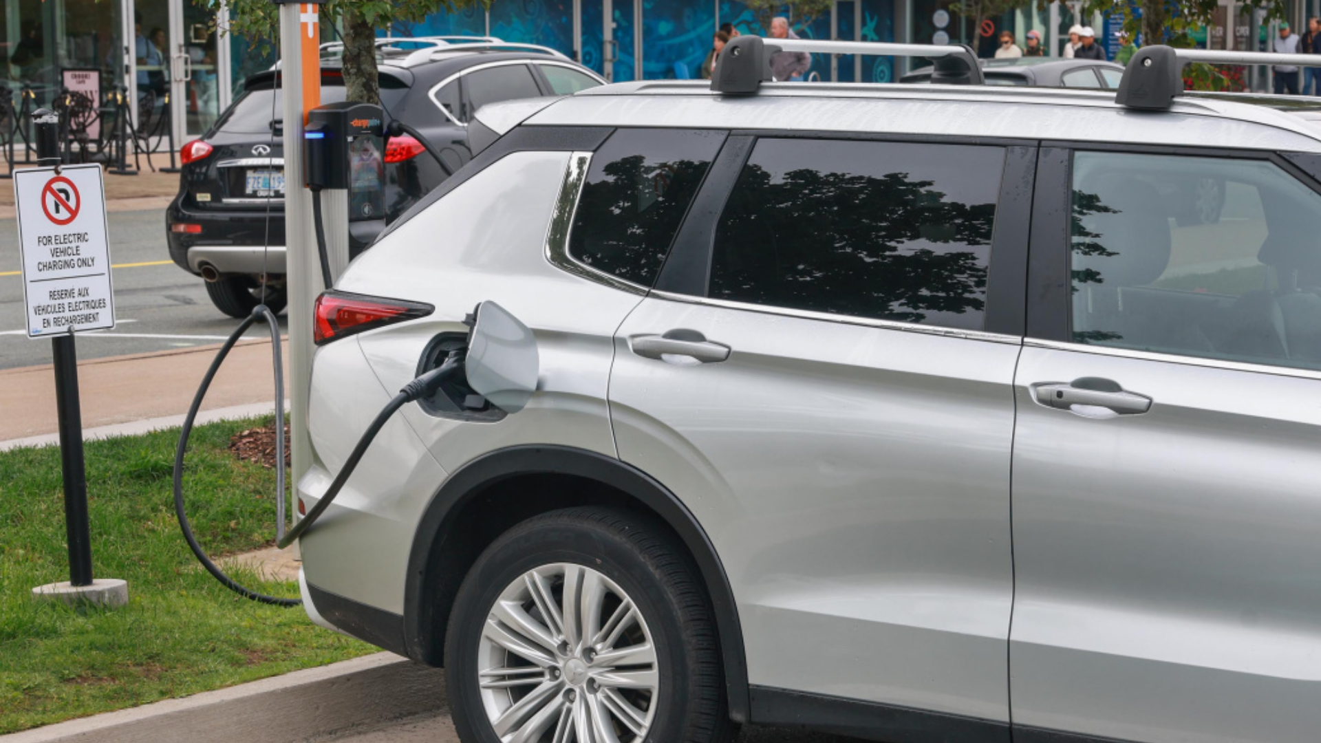 Silver electric vehicle plugged into public charging station on urban street with signage and pedestrians