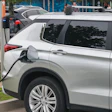 Silver electric vehicle plugged into public charging station on urban street with signage and pedestrians
