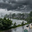 Rainy coastal road with storm clouds, residential buildings, and bridge visible across turbulent water