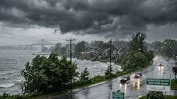Rainy coastal road with storm clouds, residential buildings, and bridge visible across turbulent water