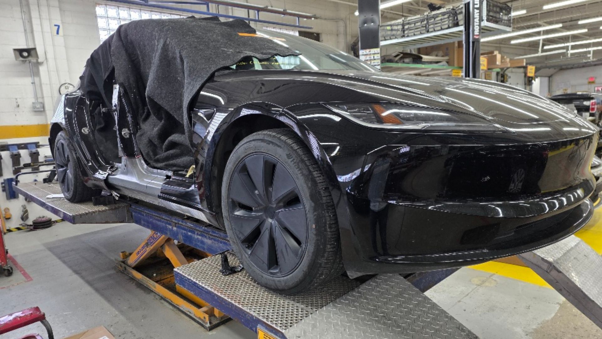Black electric vehicle on service lift in factory workshop with open door and technician overhead