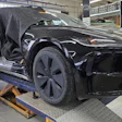 Black electric vehicle on service lift in factory workshop with open door and technician overhead