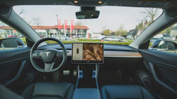 Tesla vehicle interior dashboard view with steering wheel and touchscreen display facing a shopping center parking lot