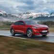 Red Ford Mustang Mach-E electric vehicle driving on wet road with snow-capped mountains in background